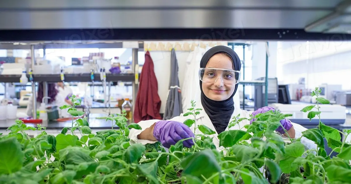 A smiling researcher in safety glasses and gloves checks rows of green plants in a university lab
