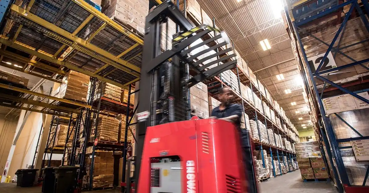 A worker driving a forklift down a warehouse aisle stacked high with boxed food on pallets