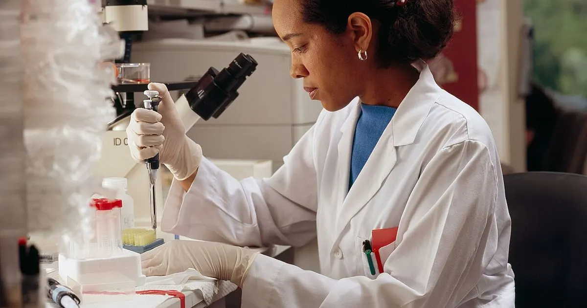 A researcher in a white lab coat and gloves uses a pipette at a laboratory bench (US National Cancer Institute archive photo)