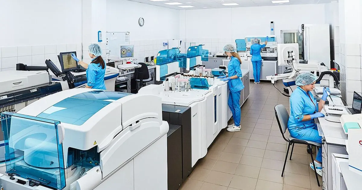 Laboratory staff in blue gowns and caps working at analysers and computers in a bright clinical lab
