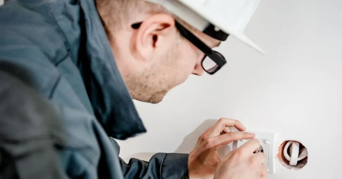 An electrician in a white hard hat and glasses fitting a wall socket