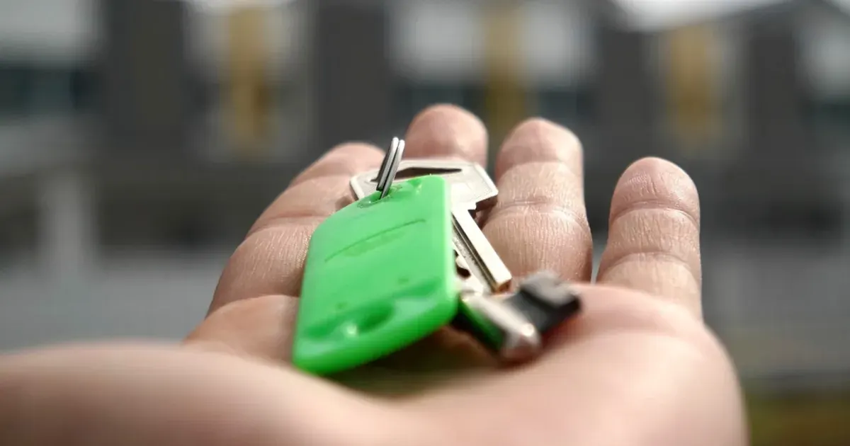 A hand holding a set of house keys on a green key tag in front of a blurred house